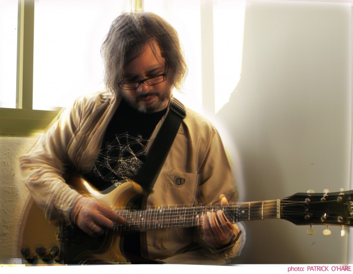 Sean Moran playing the guitar with a window behind him