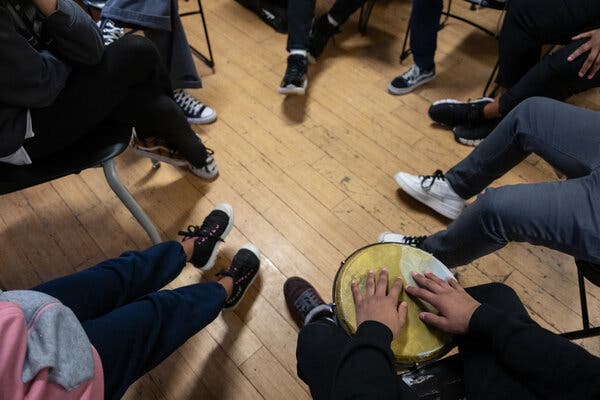 A view overhead of eight children sitting in a tight circle; only their legs and sneakers are visible. One has his hands on an African drum.