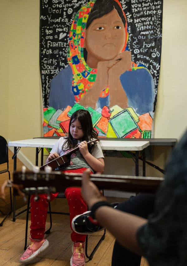 A young girl sits in a chair, intensely focused on a ukulele. Behind her is a large mural of a young girl wearing a head scarf.