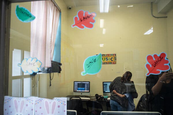 A view through a window into a classroom where a teacher hugs a student. Leaf-shaped stickers are attached to the glass, reading: “Together,” “Safe,” “Laugh,” “Sing,” “Dance.”